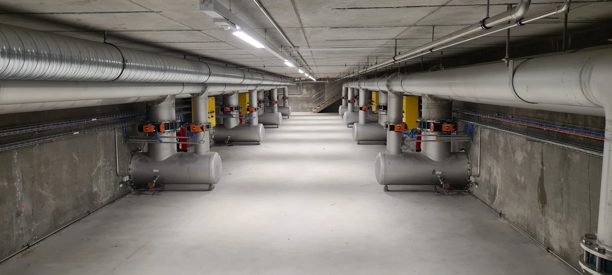 Eine betonierte Halle mit langen Rohren links und rechts. Dazwischen jeweils gelbe Pneumatikventilschränke.